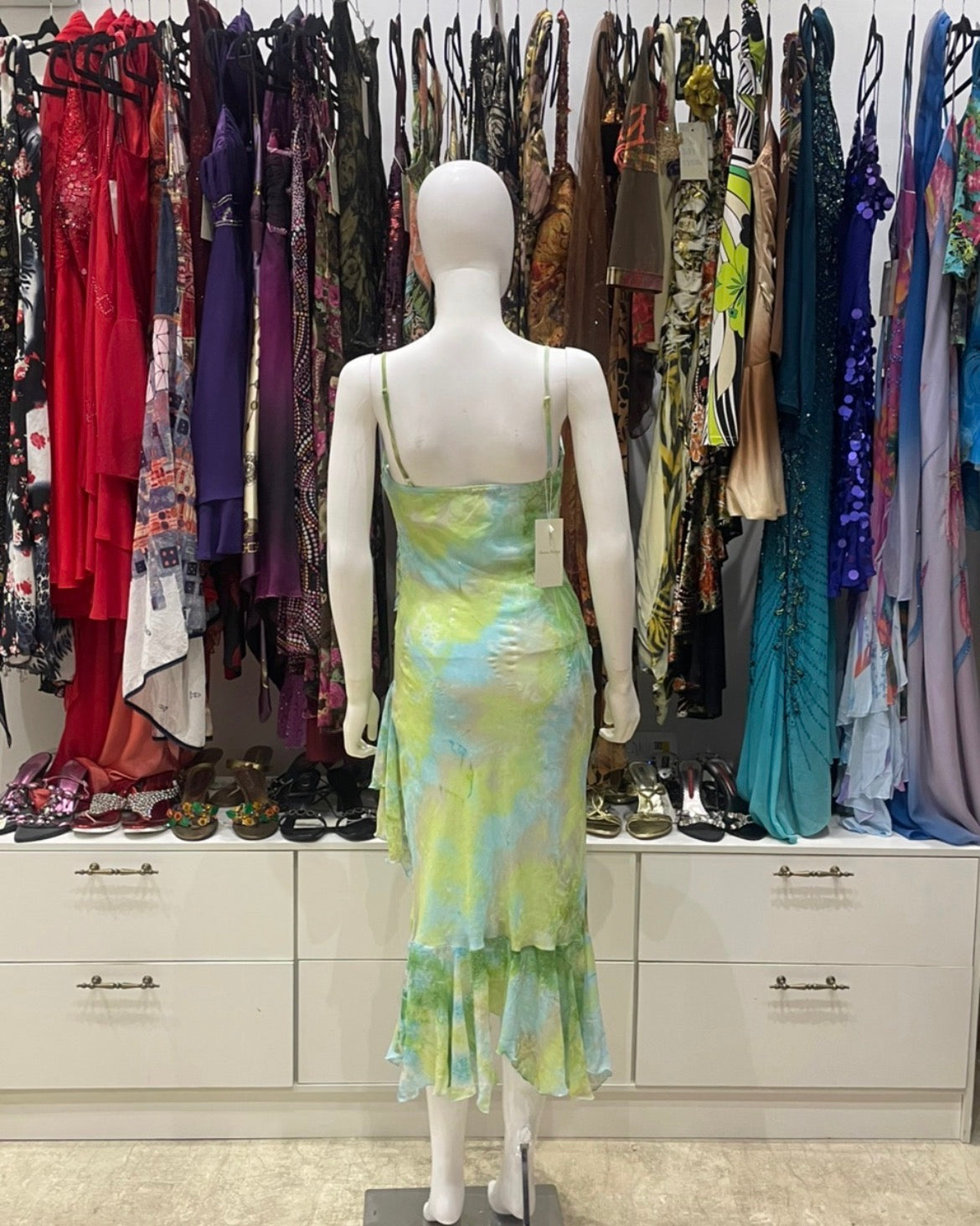 A sleeveless ruffled midi vintage dress in a yellow, green, and blue tie-dye pattern, displayed on a white mannequin from the back in a clothing store.
